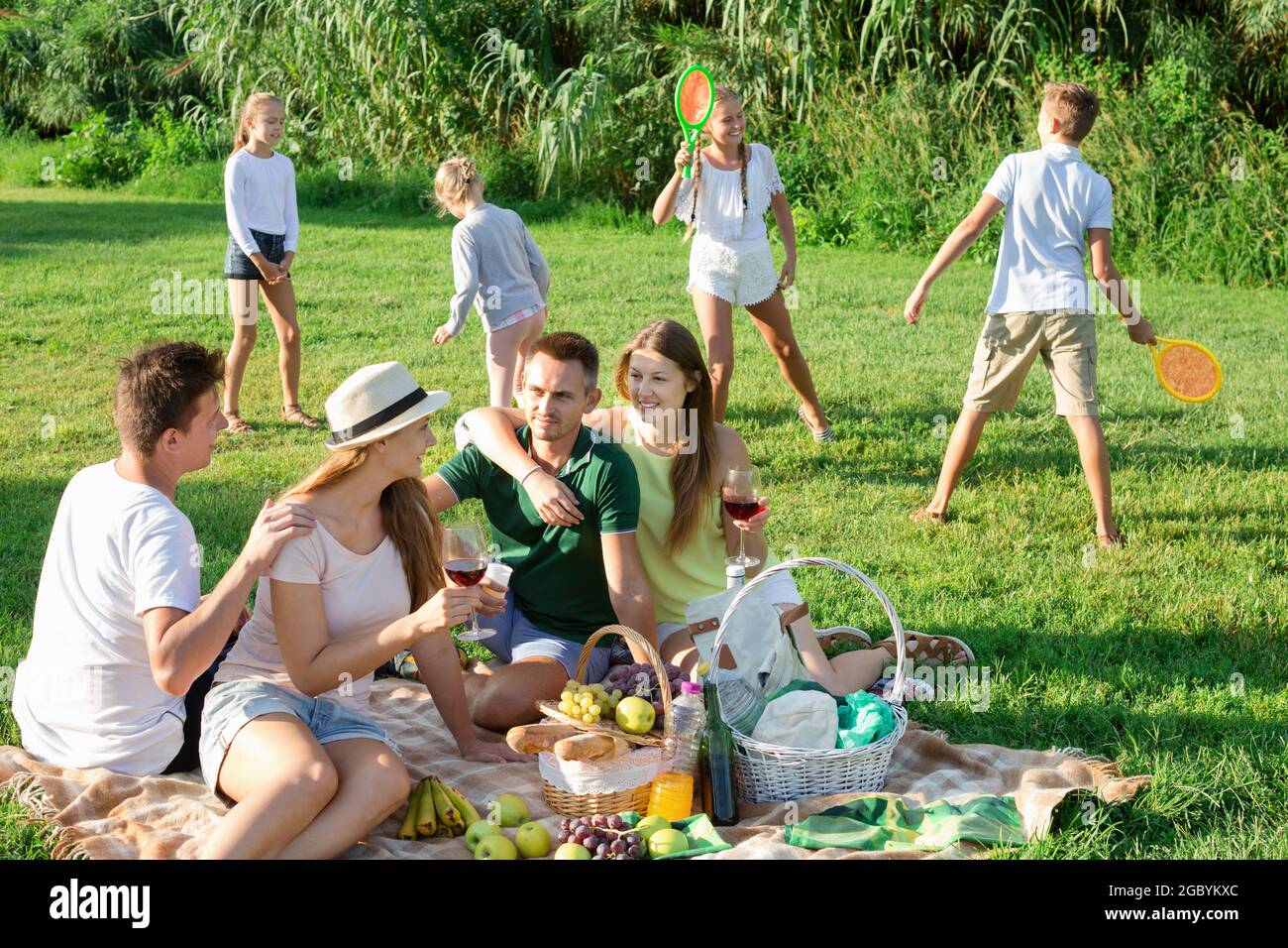 Frauen und Männer picknicken im Sommerpark mit glücklichen Kindern, die dahinter spielen Stockfoto