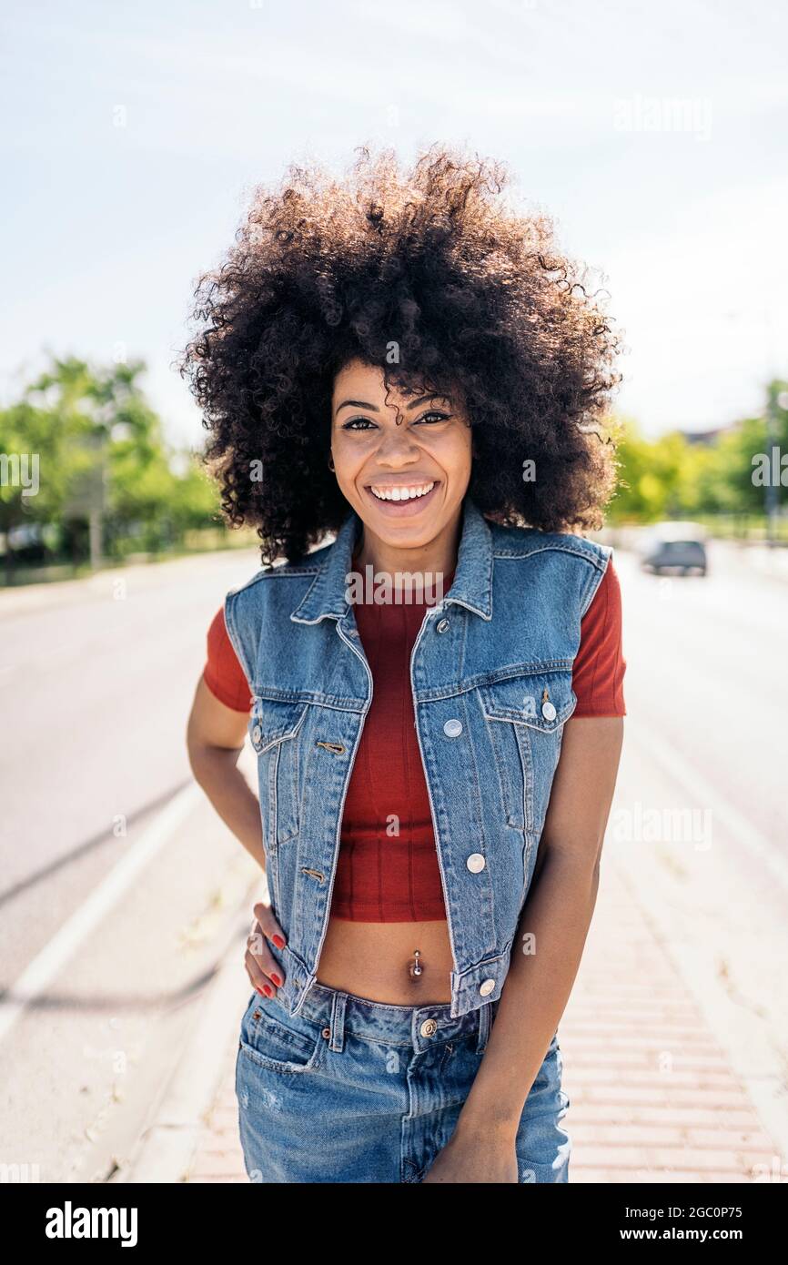 Fröhliche schwarze Frau mit kühlen Afro-Haaren lächelt und schaut auf die Kamera auf der Straße. Stockfoto