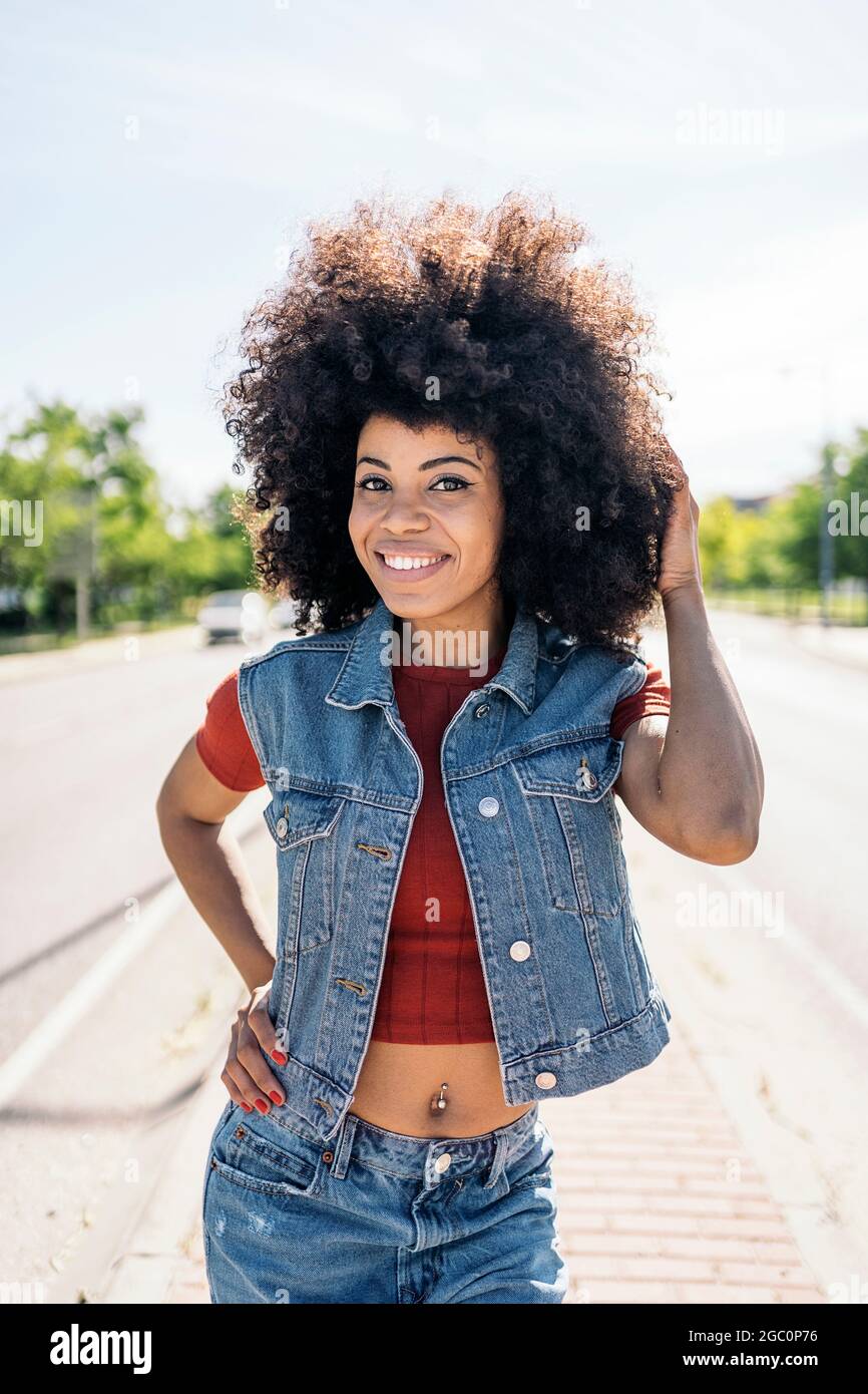 Fröhliche schwarze Frau mit kühlen Afro-Haaren lächelt und schaut auf die Kamera auf der Straße. Stockfoto