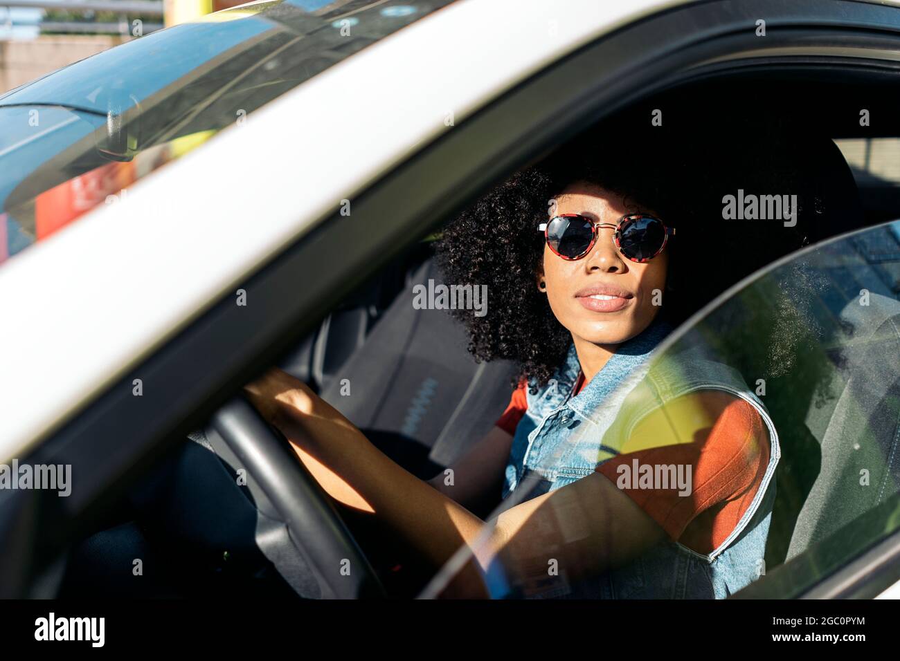 Coole Frau mit Afro-Haaren, die ihr Auto fährt und die Kamera anschaut. Sie trägt eine Sonnenbrille. Stockfoto