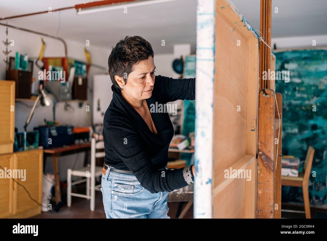 Fokussierte Malerin, die sich in ihrer Garage mit blauer Farbe auf einer großen Leinwand entspannt. Stockfoto