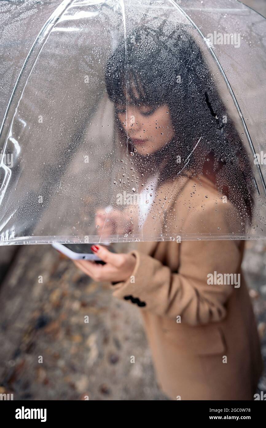 Unerkannte junge Frau mit Regenschirm, die ihr Mobiltelefon an regnerischen Tagen auf der Straße benutzte. Stockfoto