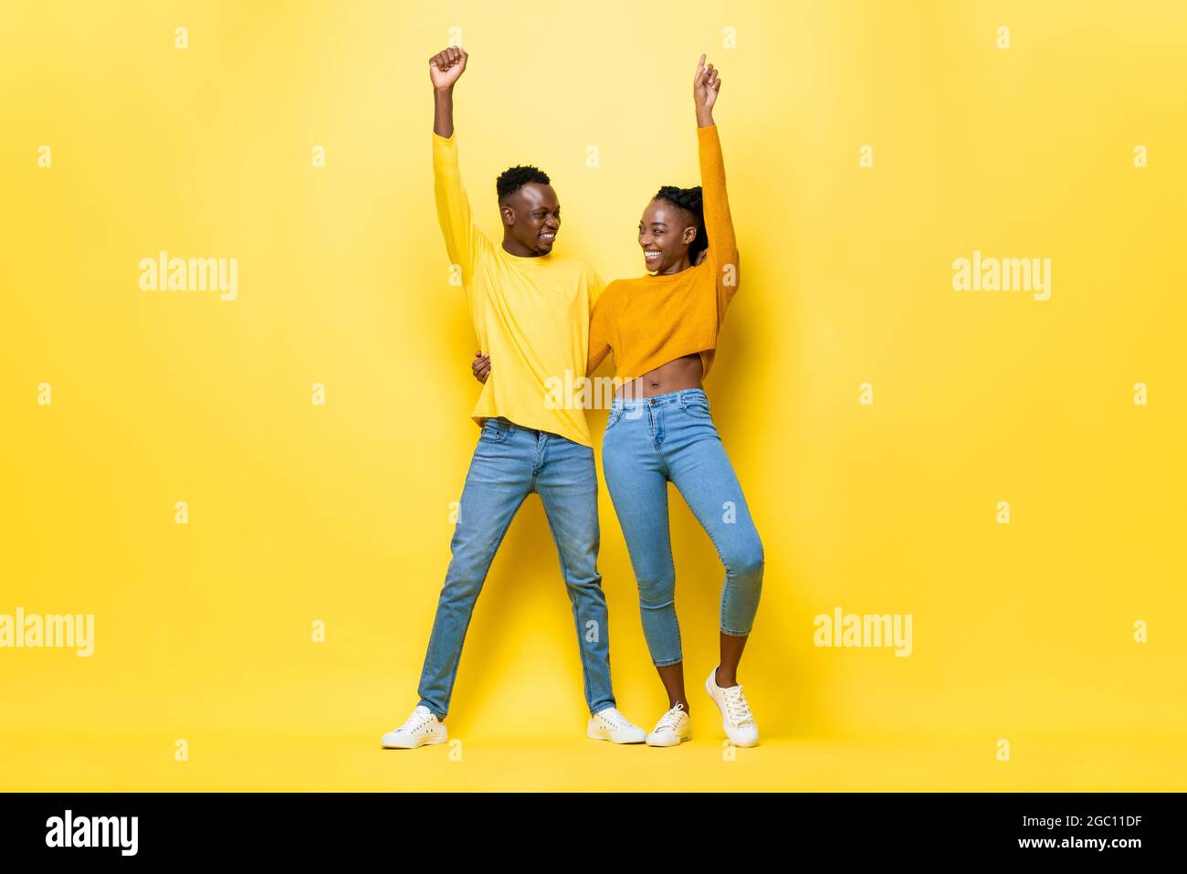Ganzkörperportrait eines glücklichen lächelnden jungen afrikanischen Paares, das die Arme hochhebt und sich gegenseitig in einem gelben, isolierten Studiohintergrund hält und schaut Stockfoto