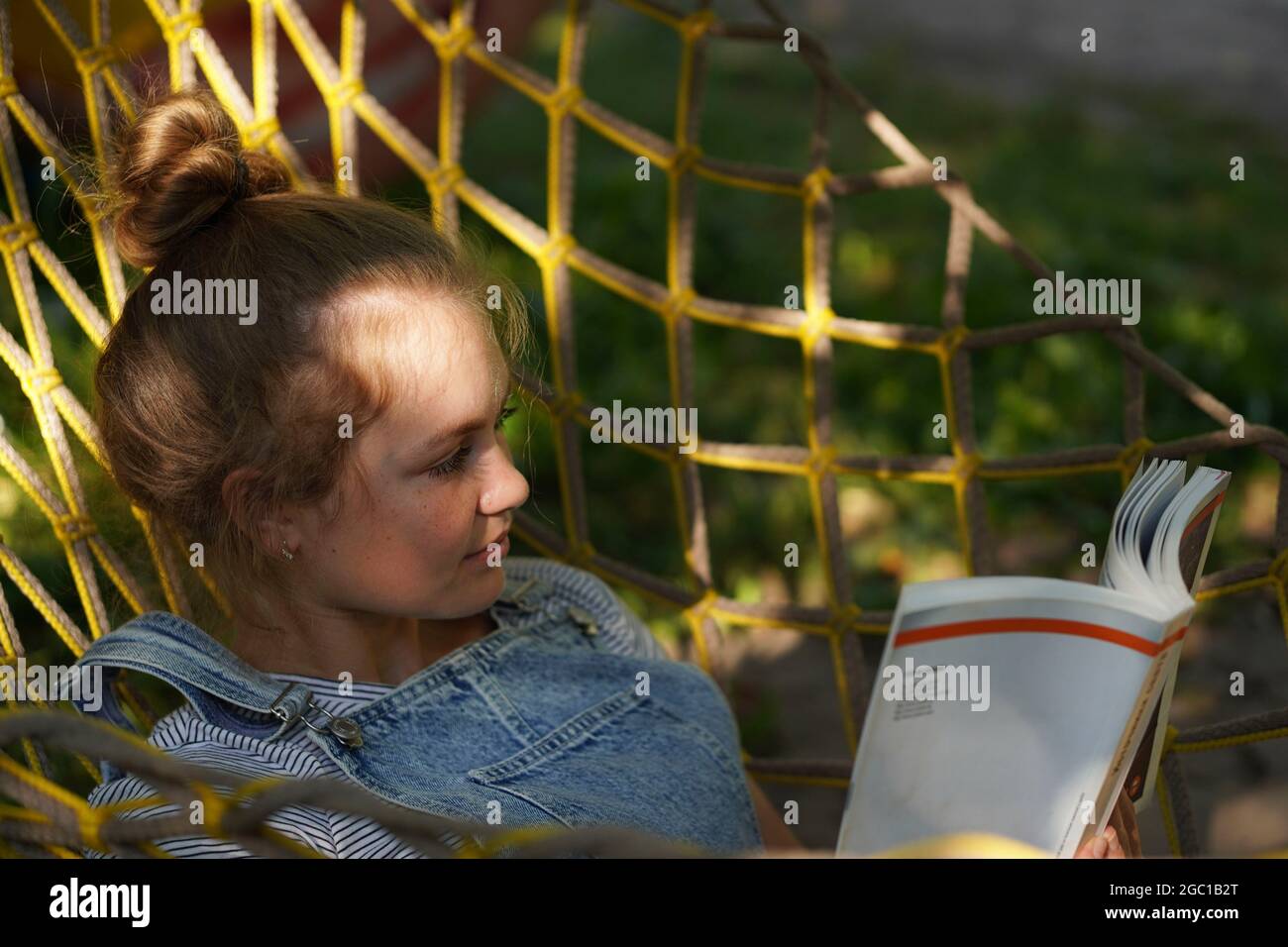 Junge blonde Studentin, die lächelt und ein Buch liest, während sie auf EINER Hängematte im Garten liegt. Frau, die sich in der Hängematte entspannt. Zurück zur Schule Universität College Stockfoto