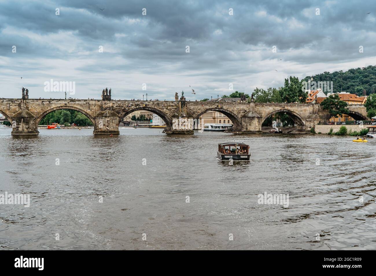 Prag, Tschechische Republik - August 4,2021. Kleine hölzerne Fähre Boot mit Passagieren überqueren Moldau, Karlsbrücke im Hintergrund. Lokalen Wassertransport Stockfoto