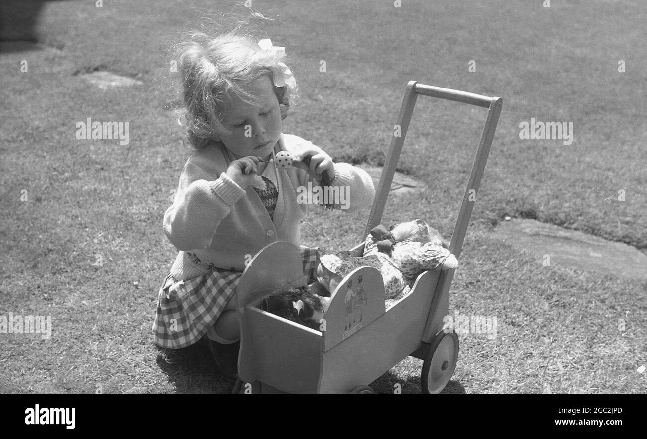 1960, historisch, draußen in einem Garten, knieend auf dem Gras, ein junges Mädchen mit ihrem kleinen Holzpuppen-Kinderwagen, voll von weichen Spielzeugen, darunter ein Teddybär, England, Großbritannien. Stockfoto