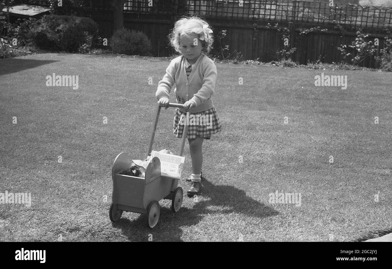 1960er Jahre, historisch, draußen in einem Garten auf dem Gras, ein junges Mädchen schob ihre kleinen Holzpuppen Kinderwagen, England, Großbritannien. Stockfoto