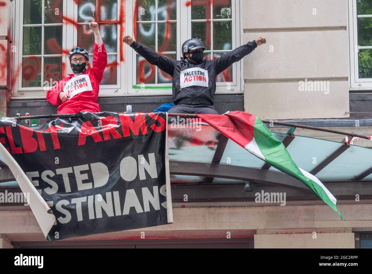 London, Großbritannien. August 2021. Zwei Demonstranten der Palestine Action sitzen mit Spruchbändern auf der Kante im ersten Stock über dem Eingang zu den Londoner Büros des israelischen Waffenherstellers Elbit Systems in Kingsway, mit Spruchbändern und gemalten Botschaften "Elbit tötet", "Shut Elbit Down". Elbit liefert Waffen an die israelische Regierung, um Kriegsverbrechen in Gaza zu begehen, und vermarktet ihre Waffen als "kampferprobt" wegen ihres Einsatzes gegen Palästinenser. Andere spielten Trommeln auf der gegenüberliegenden Straßenseite, um auf den Protest aufmerksam zu machen. Peter Marshall/Alamy Live News Stockfoto