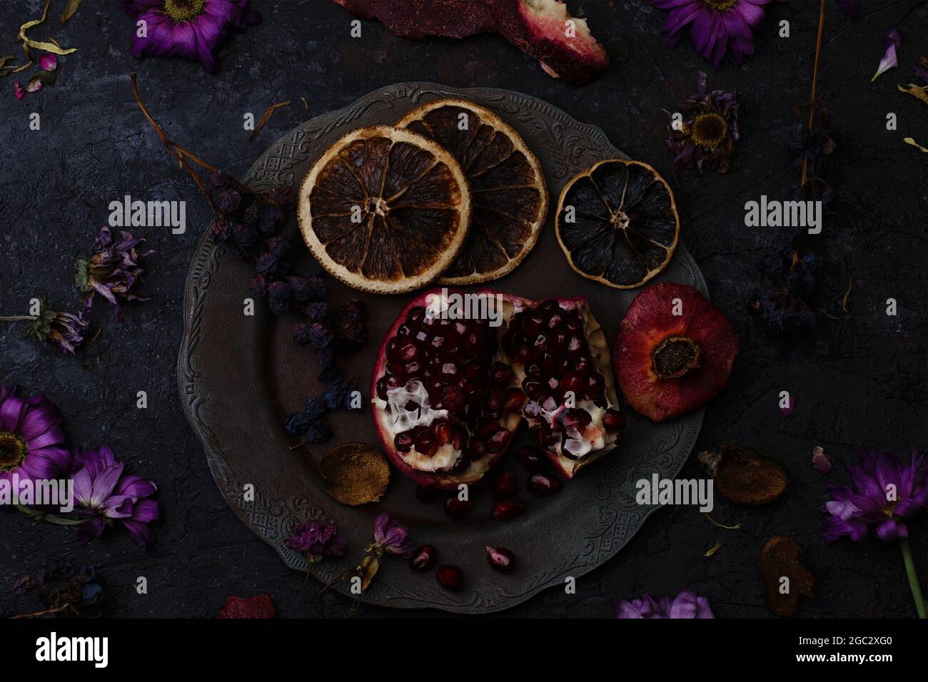 Reifer Granatapfel in einem Vintage-Teller, ein Bund trockener Trauben und orangefarbene Scheiben zwischen welkenden Blumen auf dunklem Steingrund Stockfoto