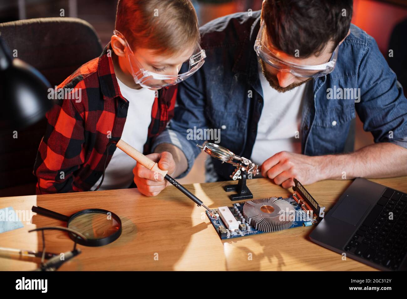 Fürsorglicher Vater mit kleinem Sohn in Freizeitkleidung und Brillen, die mit Lötkolben arbeiten, während sie gebrochene Details vom Computer reparieren. Konzept von Elternschaft und Lehre. Stockfoto