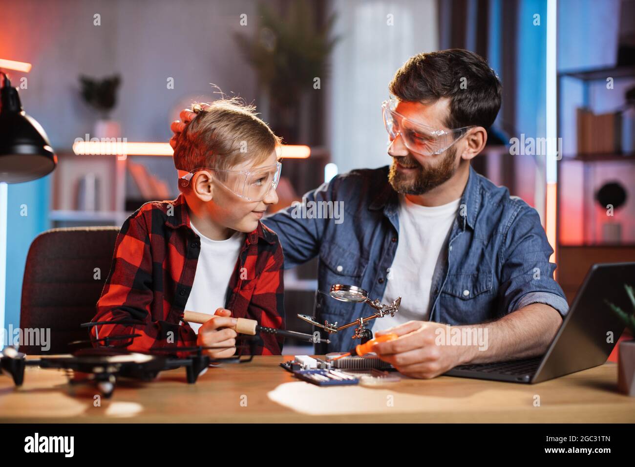 Positiver kaukasischer Vater lehrt seinen kleinen Sohn, wie man Displaykarten von einem modernen Laptop aus repariert. Mann und Junge arbeiten zu Hause mit Lötkolben. Stockfoto