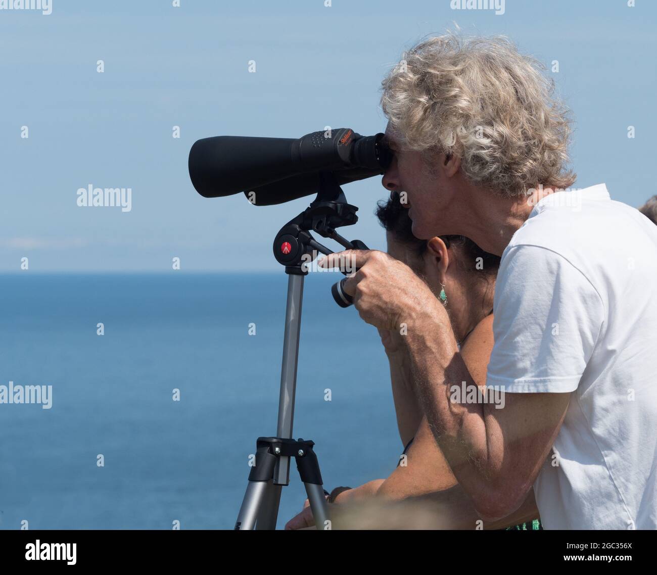 Ein Mann schaut durch ein großes Fernglas von skymaster, das auf einem Manfrotto-Stativ befestigt ist.Meer und Ozean im Hintergrund Stockfoto