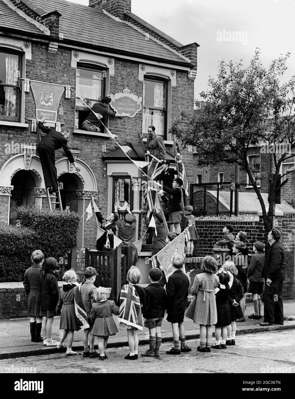 Die Coronation Road, West Ham, wird seinem Namen gerecht, während der Krönung wird jedes Haus vollständig dekoriert und 27 königliche Schilde schmücken die Straße und Feiern werden auch auf der Straße gefeiert. Eine bekannte Display-Organisation stellt alle Dekorationen kostenlos zur Verfügung, und heute ist die Coronation Road für den Verkehr gesperrt, damit die Bewohner ihre Häuser dekorieren können. Das Foto zeigt die Bewohner eines der Häuser der Coronation Road, die anfangen, die Dekorationen zu bauen. Mai 1953 Stockfoto