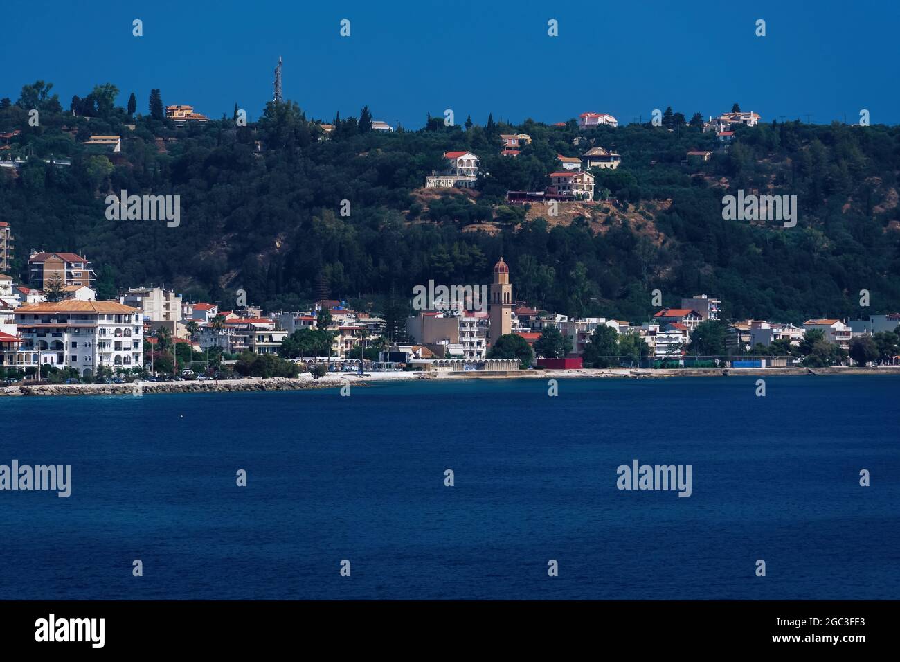 Blick auf Zakynthos, die Hauptstadt der griechischen Insel, mit flachen Gebäuden am Wasser. Landschaft der orthodoxen Kirche um rote Ziegelhäuser neben ruhigen Meer & Hügel. Stockfoto