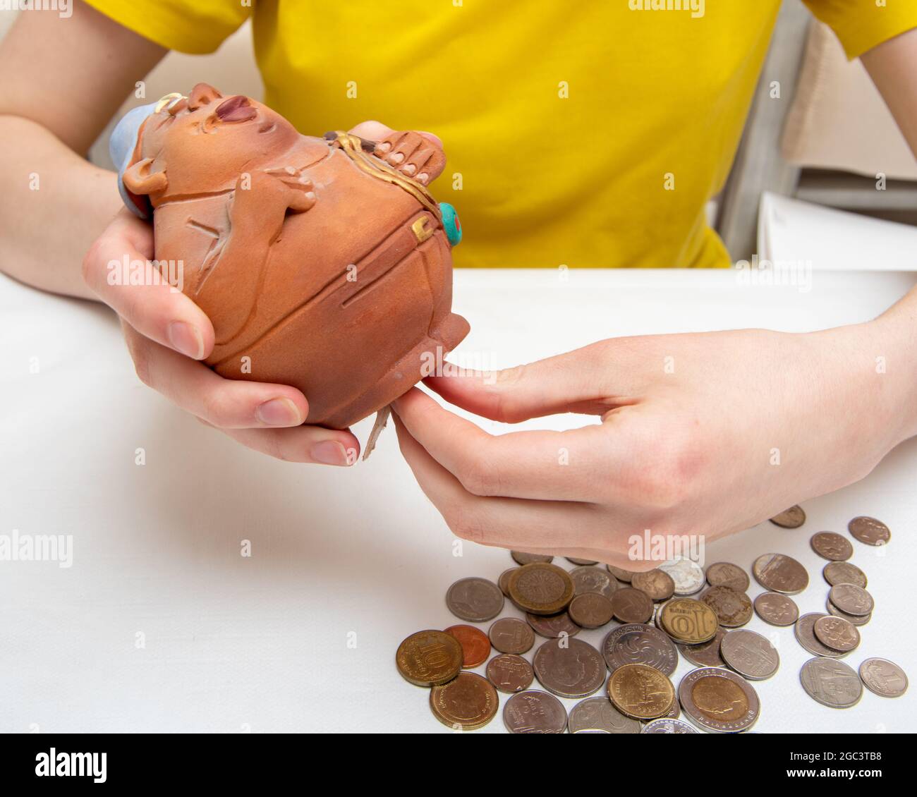 Geld sparen. Die Hand zieht Münzen heraus. Brauchen Sie ein offenes Sparschwein mit Münzen, verstreuten Münzen verschiedener Länder, auf weißem Hintergrund, Nahaufnahme Stockfoto