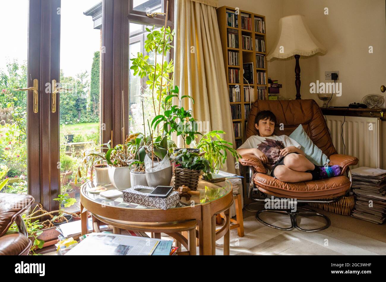 Ein Junge entspannt sich in einem bequemen Stuhl mit einem Glastisch, der mit Zimmerpflanzen bedeckt ist. Eine heimelige Lifestyle-Szene aus einem Familienhaus. Stockfoto