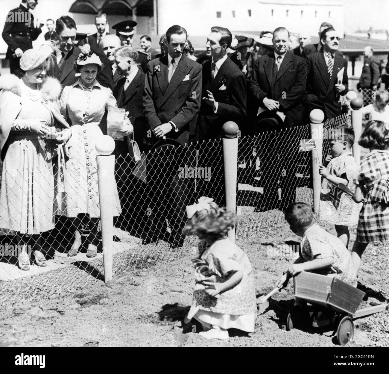 Diese Kinder spielten glücklich in Castlereagh, Belfast, als die Königin und Prinzessin Margaret heute (Freitag) während ihrer Nordirland-Tour über den Draht guckerten. Die Queen trägt ein pulverblaues Seidenkleid mit passendem Cape. Prinzessin Margaret ist in Austerngrau Kordelseidenmantel über einem rosa Seidenkleid . Ihr passender Hut ist aus rosa Stroh mit einer Kakade. Juni 1951 Stockfoto