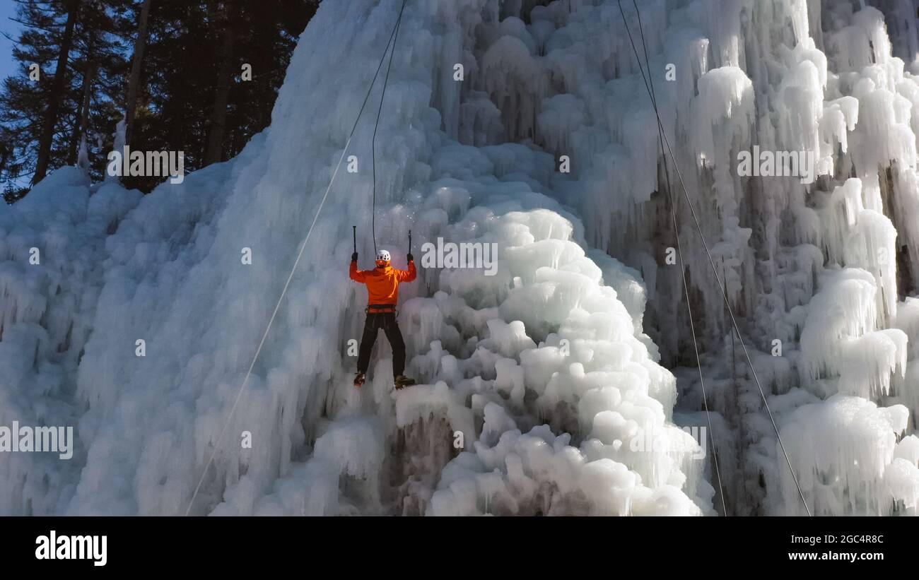 Luftaufnahme eines gefrorenen Eiswasserfalls, und der männliche Kletterer schreitet den Hang hinauf, während ein Seil, das an seinem Geschirr befestigt ist, ihn daran hindert, zu fallen Stockfoto