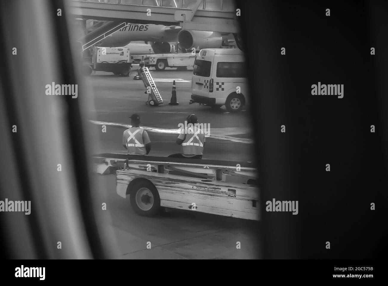 Graustufenaufnahme einer Flughafenrampe mit Bodenpersonal und Fahrzeugen aus einem Flugzeugfenster Stockfoto