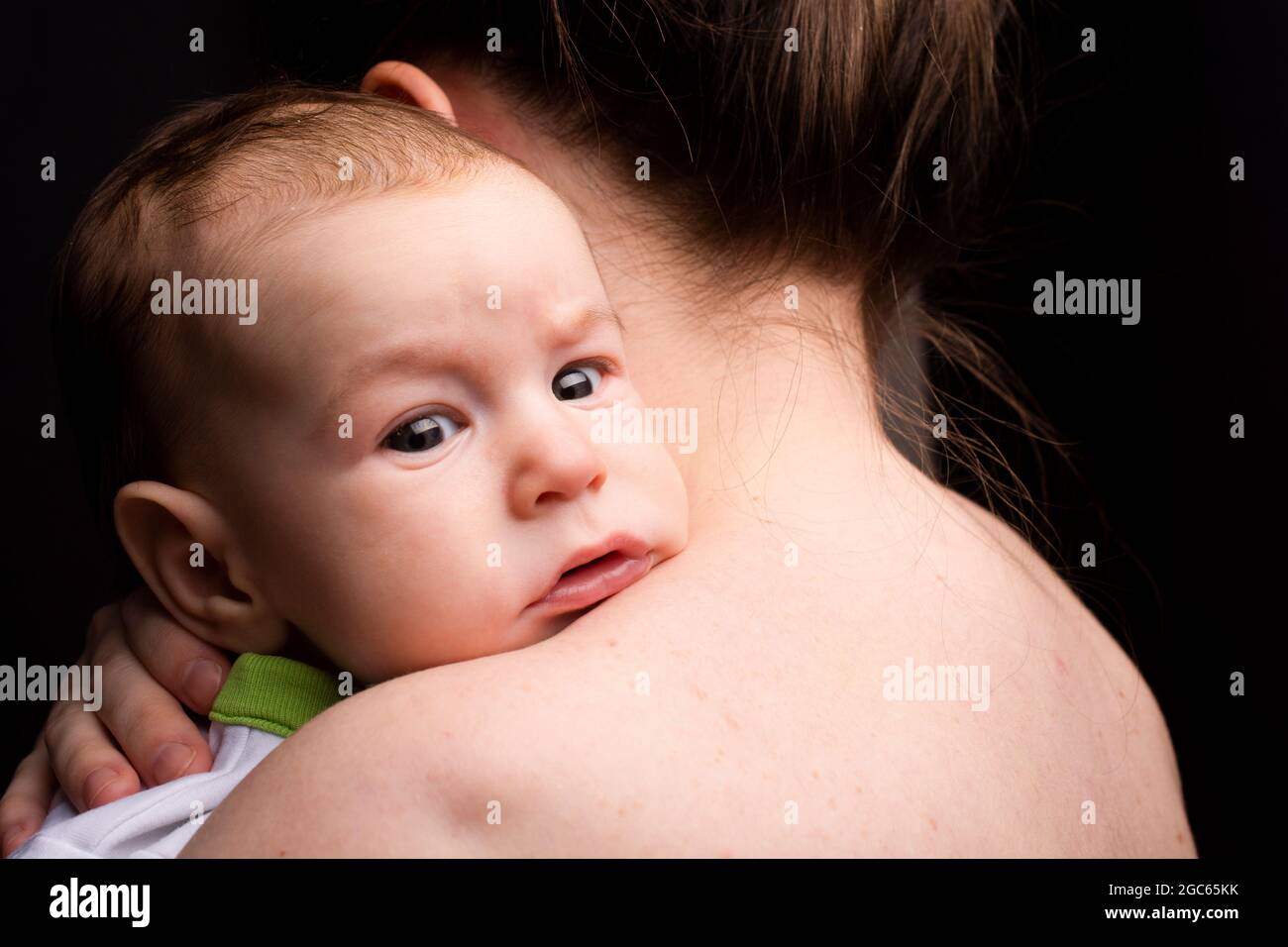 Baby auf der Schulter der Mutter, Blick auf die Kamera, auf schwarzem Hintergrund. Stockfoto