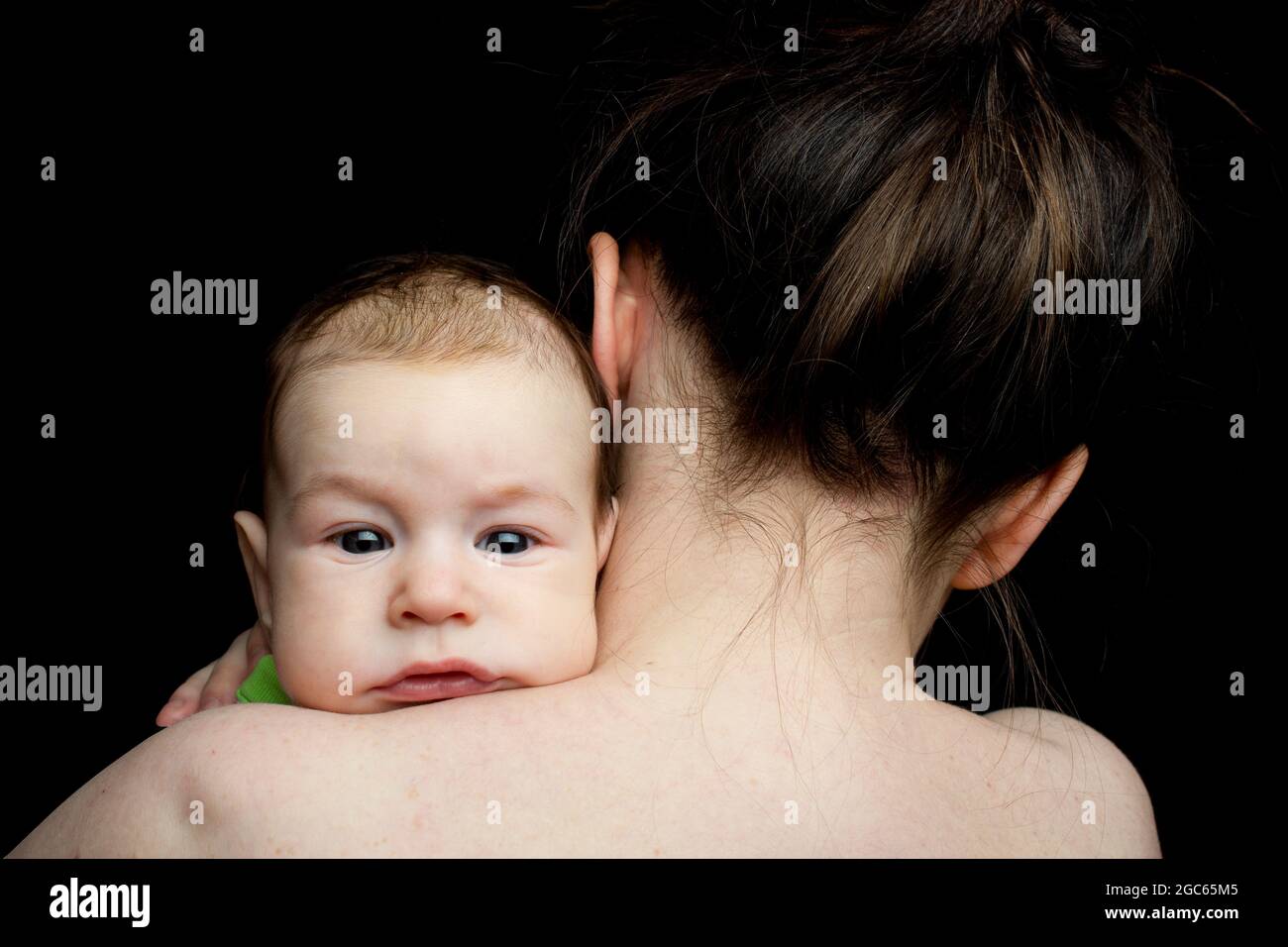 Baby auf der Schulter der Mutter, ohne Emotionen auf die Kamera schauend. Auf schwarzem Hintergrund. Stockfoto