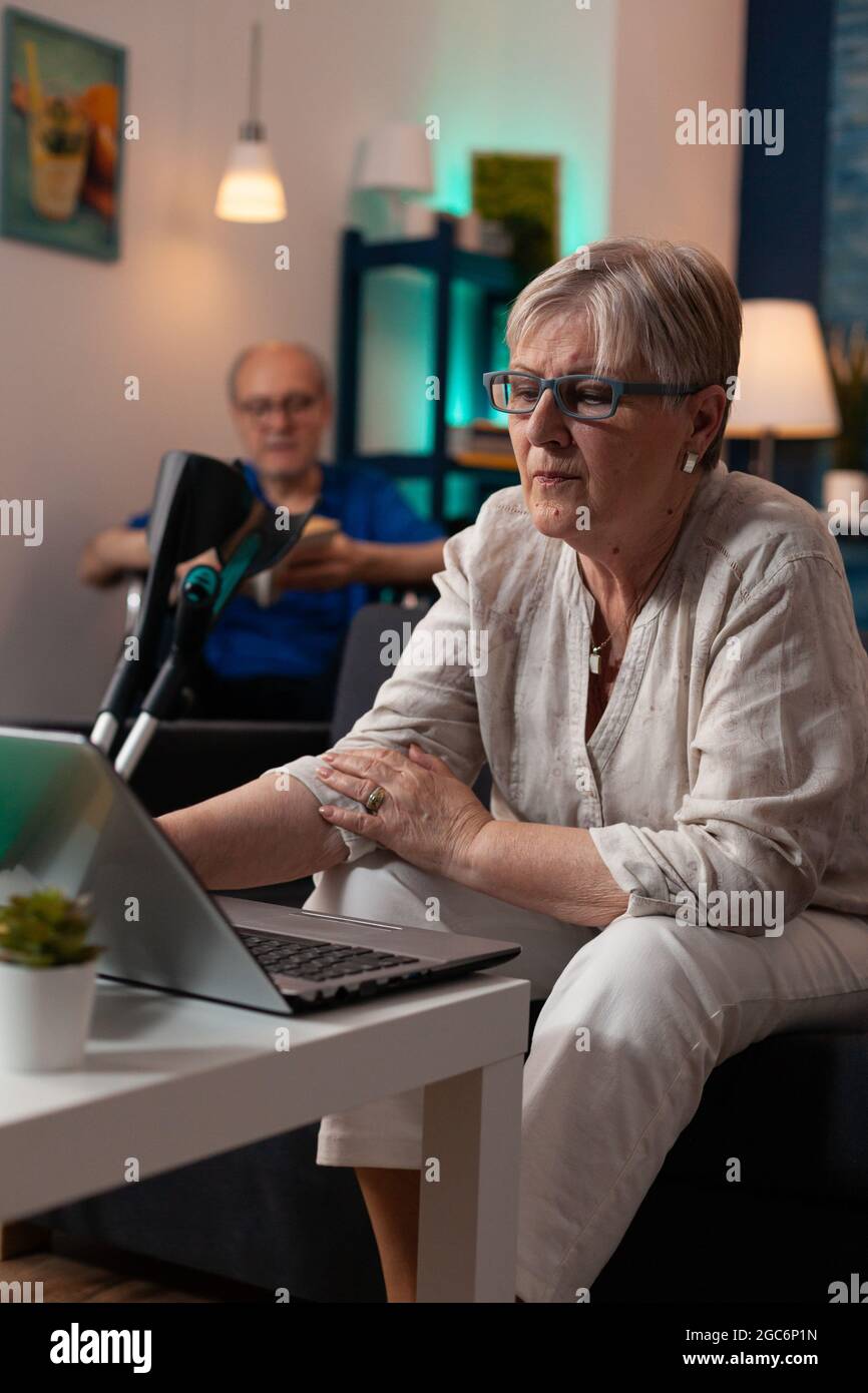 Ältere kaukasische Menschen genießen Aktivitäten zu Hause. Die alte Frau benutzt einen modernen Laptop-Computer, der auf der Couch sitzt, während der Mann im Rollstuhl liest. Ältere ältere behinderte Paare Stockfoto