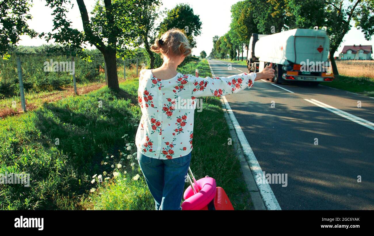 Reisende Frau hitchwandering auf einer sonnigen Straße und zu Fuß. Junge glückliche Backpacker Frau auf der Suche nach einer Fahrt, um eine Reise auf einer sonnig beleuchteten Landstraße zu beginnen. Stockfoto