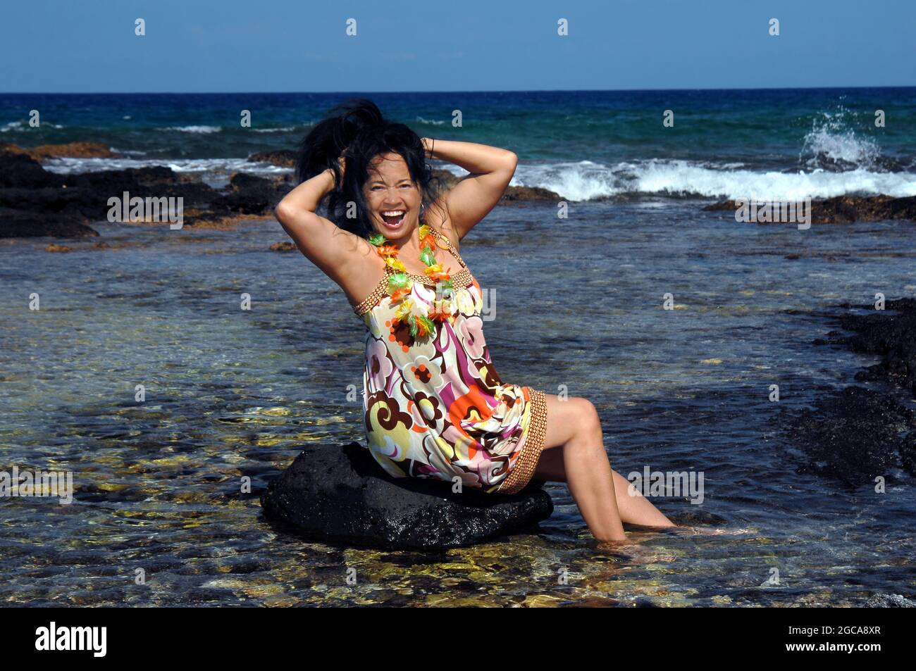 Hawaiianerin sitzt auf einem Felsen im flachen Wasser und feiert, indem sie ihre Haare hochwirft und posiert. Anaehomalu Bay an der Kohala Küste der Big Isla Stockfoto