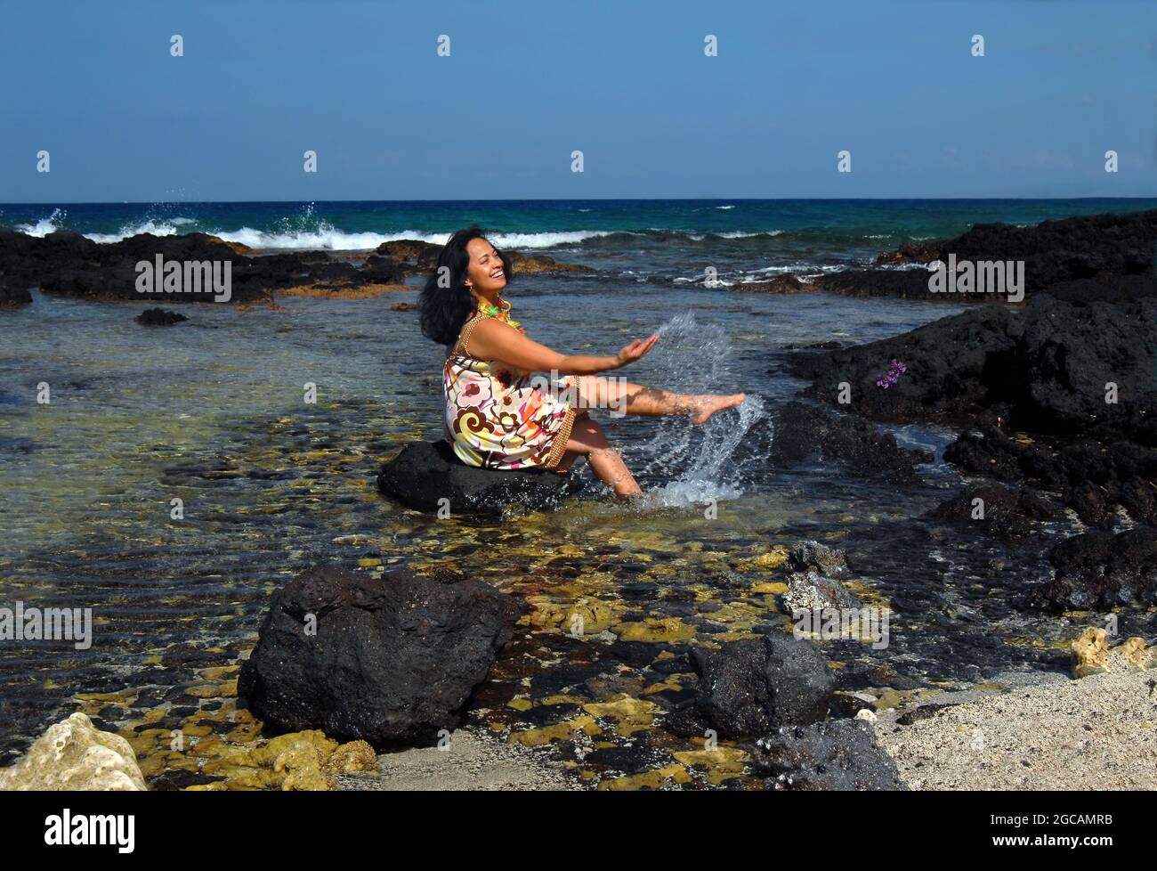 Ältere Hawaiiianerin wadet in die Untiefen und sitzt auf einem schwarzen Lavastein, der klatscht und spritzt. Stockfoto
