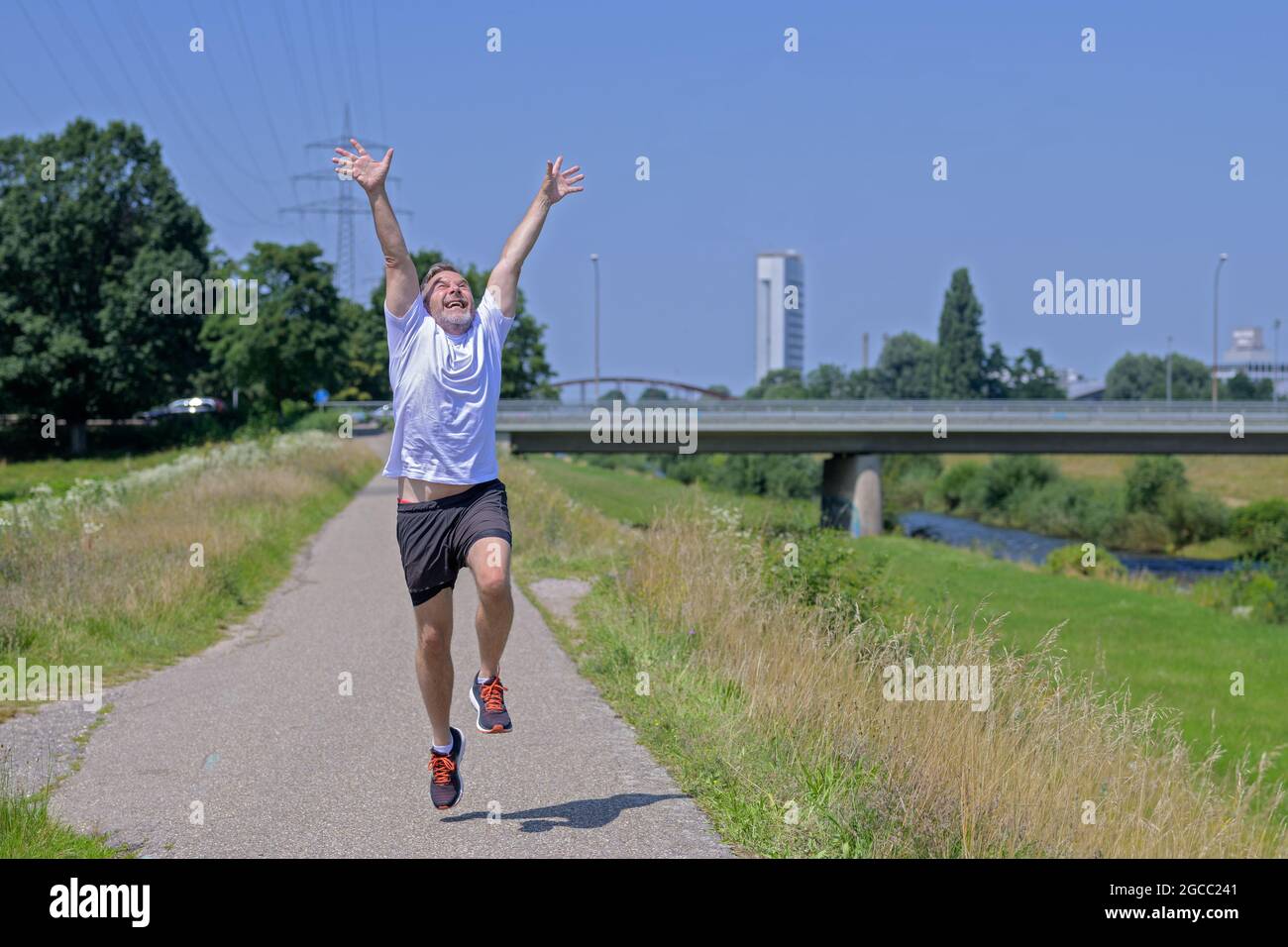 Fröhlich überschwänglichen Mann mittleren Alters joggen entlang eines Flusses feiert seine gute Gesundheit und sonniges Wetter hob seine Arme in der Jubel Stockfoto