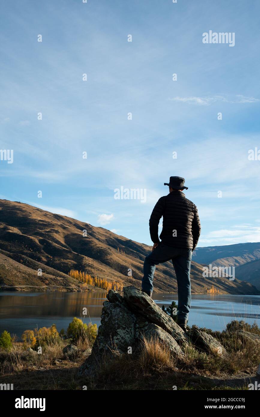 Ein Mann, der auf den Felsen am Fluss Clutha steht und die sanften Hügel im Herbst beobachtet. Central Otago, Südinsel. Vertikales Format. Stockfoto