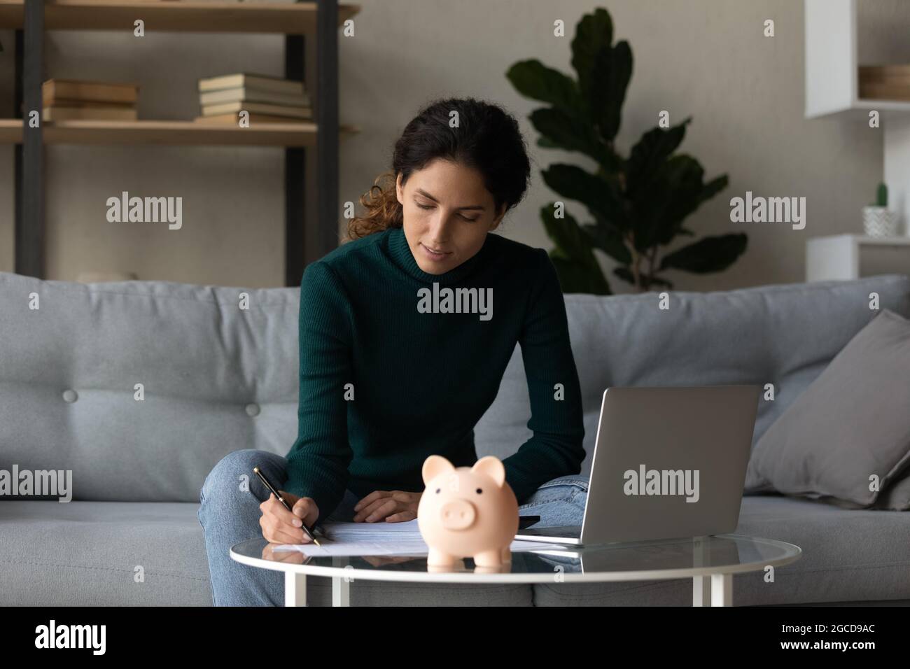 Zufriedene Frau, die Finanzen überprüft, am Tisch mit Sparschwein sitzt Stockfoto