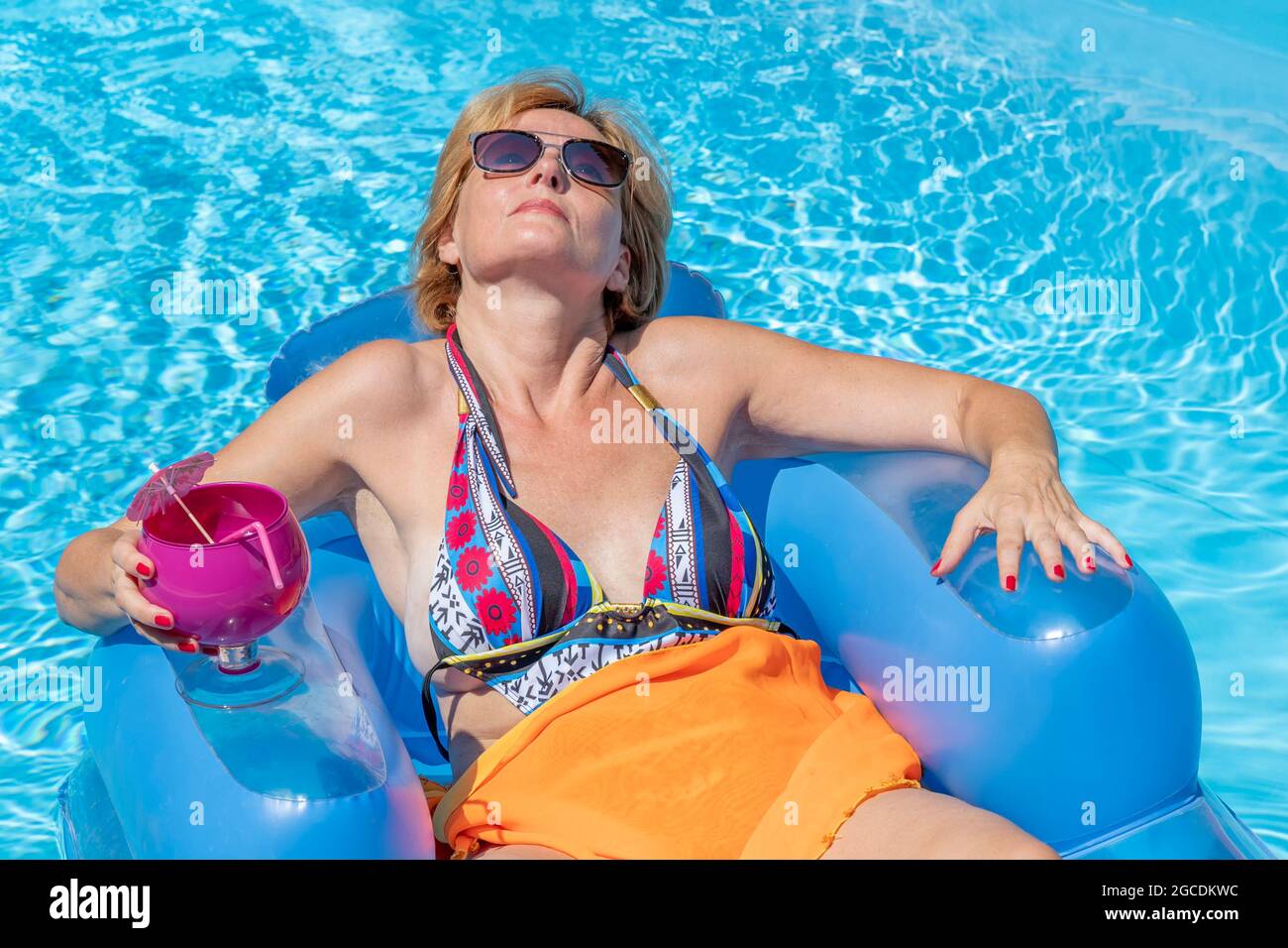 Eine weiße Frau mittleren Alters entspannt sich auf einer aufblasbaren Matratze auf dem Wasser eines Schwimmbades, mit einem Glas Cocktail in der Hand Stockfoto