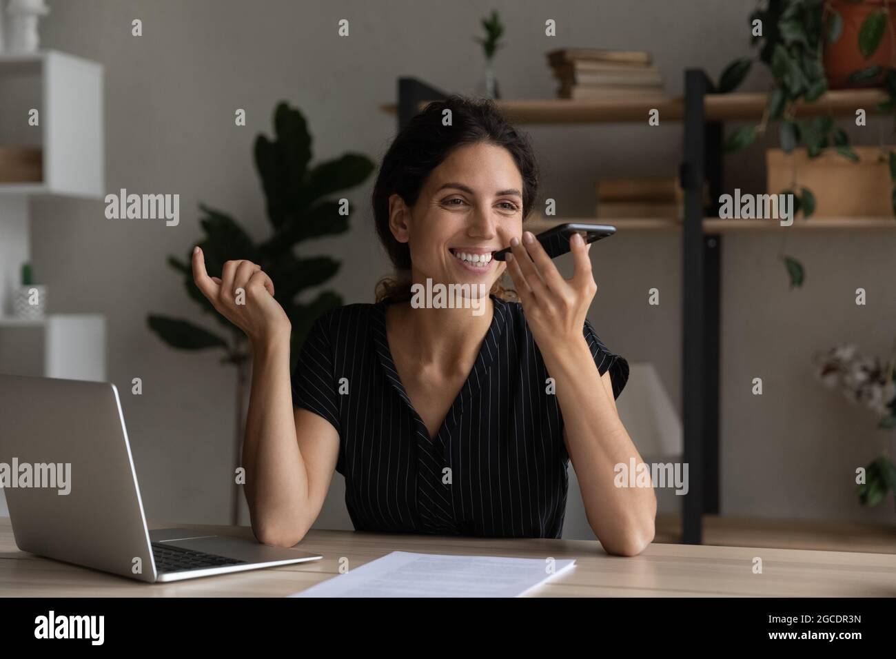 Lächelnde Geschäftsfrau, die die Sprachnachricht auf dem Smartphone am Schreibtisch aufzeichnet Stockfoto