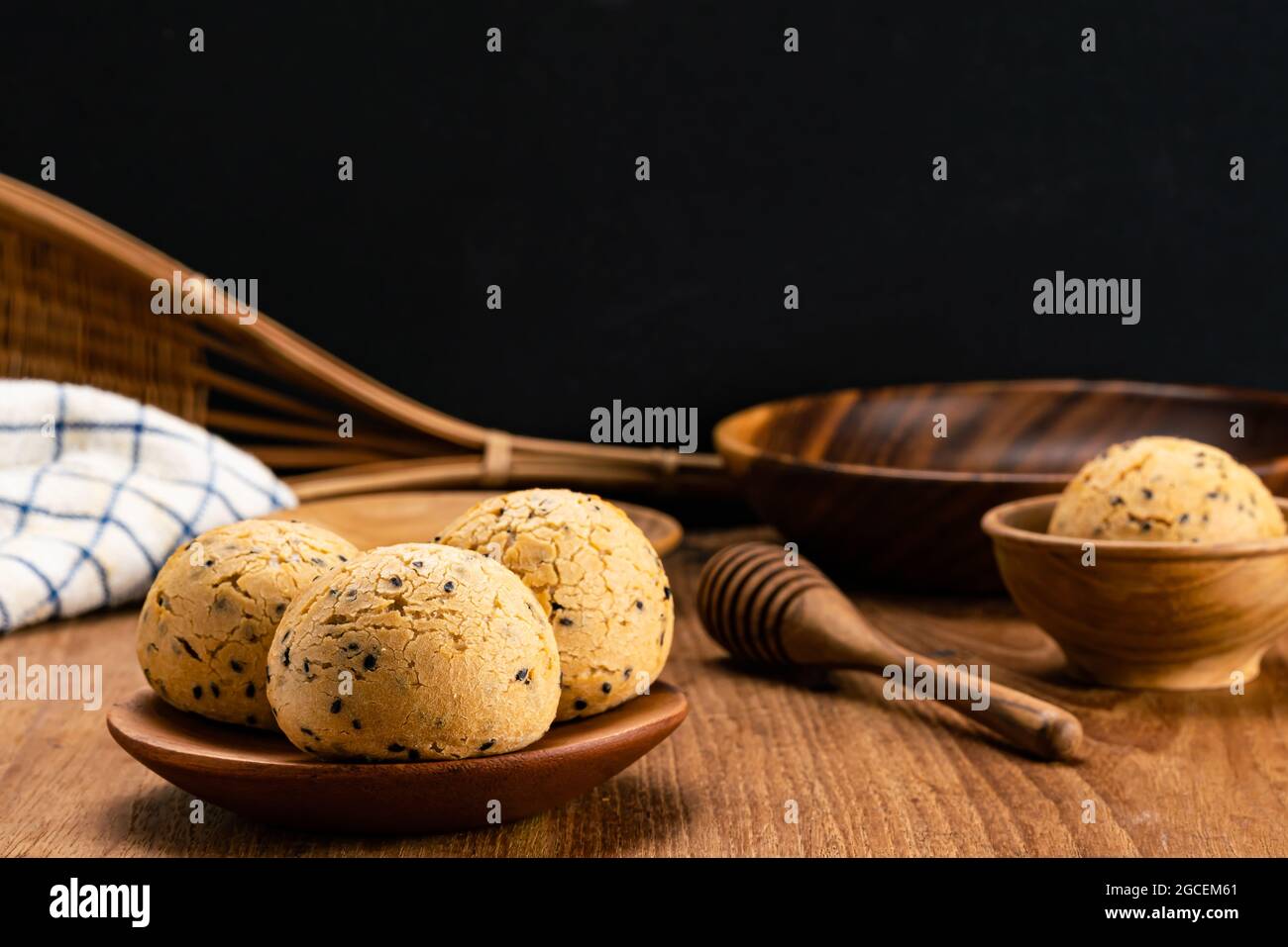 Hausgemachtes leckeres Sesam-Mochi-Brötchen in Holzplatte und in Holzschale mit Honigtaucherchen, Geschirrtuch, Bambuskorb und Holzschale auf einem Tisch. Stockfoto