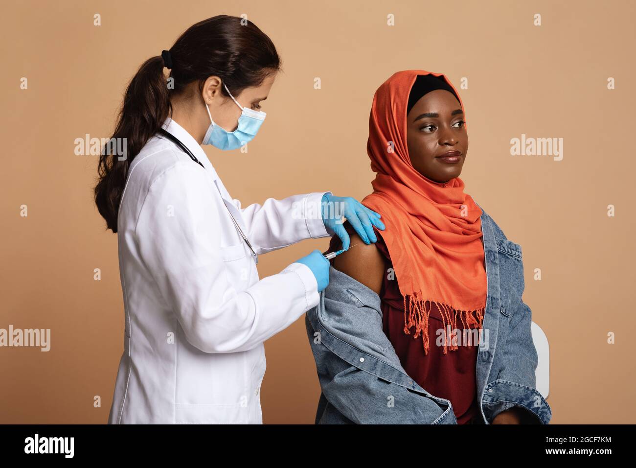 Die tausendjährige muslimische schwarze Frau im Hijab wird geimpft, sitzt auf einem Stuhl, während eine Ärztin in medizinischer Arbeitskleidung einen Impfstoff gegen Coronavi injiziert Stockfoto