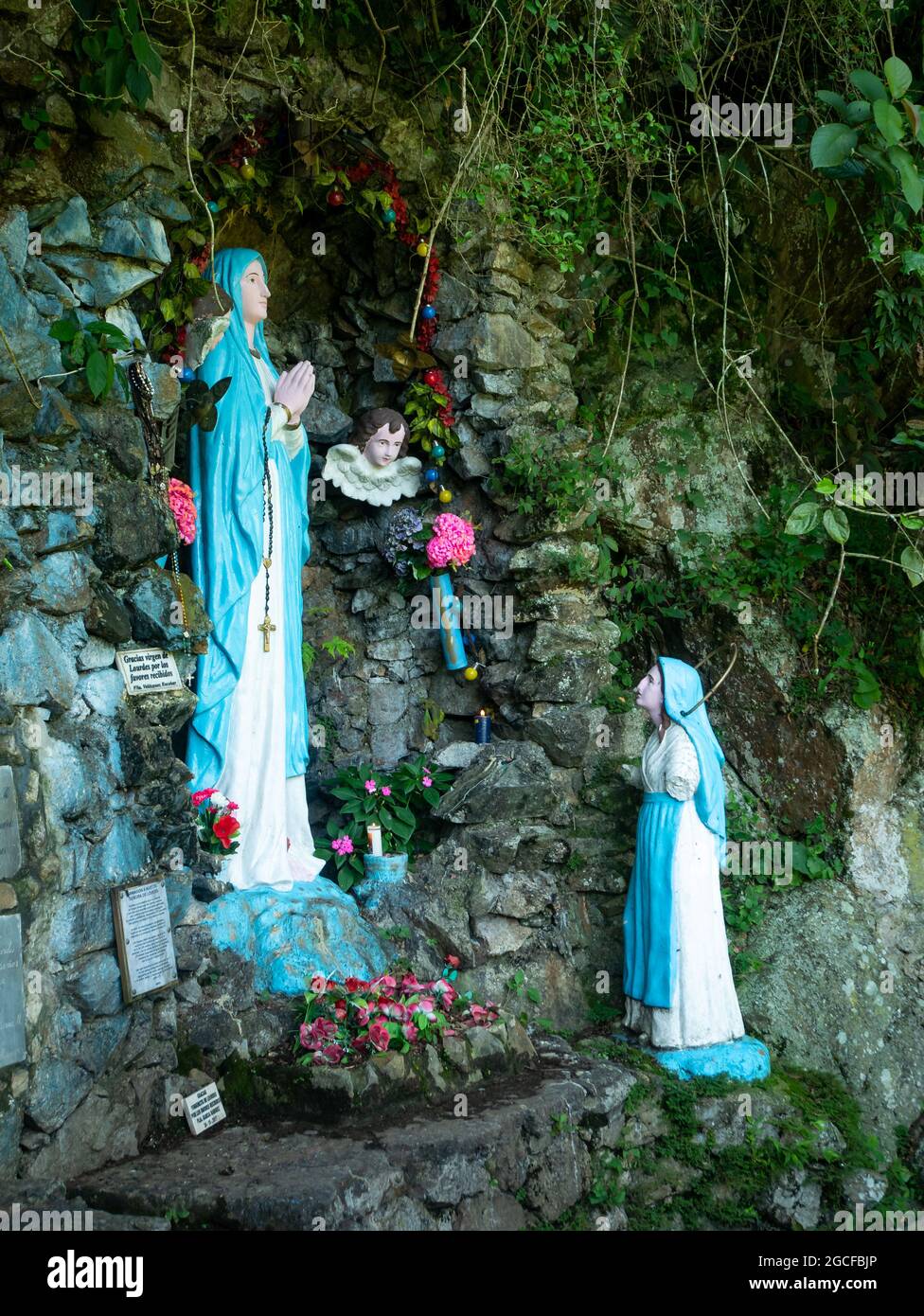 Amaga, Antioquia, Kolumbien - Juli 18 2021: Die Statuette der Jungfrau von Lourdes umgeben von Blumen und Dankesbotschaften mit einem weiteren gebrochenen Sta Stockfoto