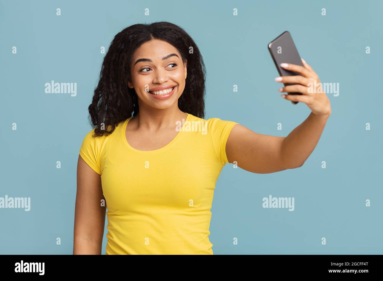 Positive schwarze Dame, die Selfie auf dem Smartphone macht, posiert und Spaß auf blauem Studiohintergrund hat. Social Media, Blogging, mobile Anwendung für sel Stockfoto