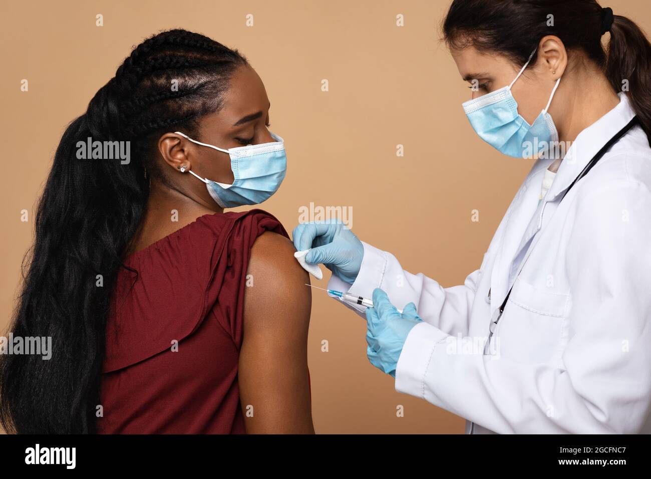 Junge Krankenschwester in medizinischer Arbeitskleidung, die für schwarze Frau Injektion gegen Coronavirus in der Schulter macht, beiger Studiohintergrund, Seitenansicht. Massenimmunisierung Stockfoto