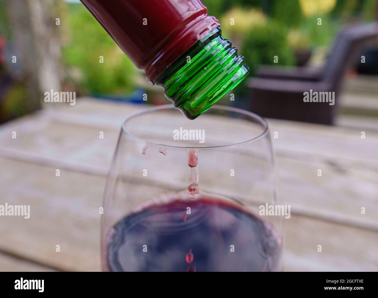 Den letzten Tropfen Rotwein aus der Flasche auf einen Glastisch aus Holz und eine verschwommene Gartenlandschaft gießen Stockfoto