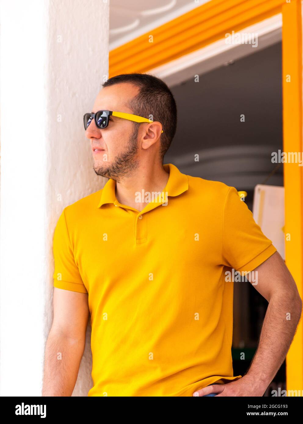 Vertikale Aufnahme eines jungen Mannes mit gelbem T-Shirt und Sonnenbrille, der in der Nähe einer Wand steht. Stockfoto