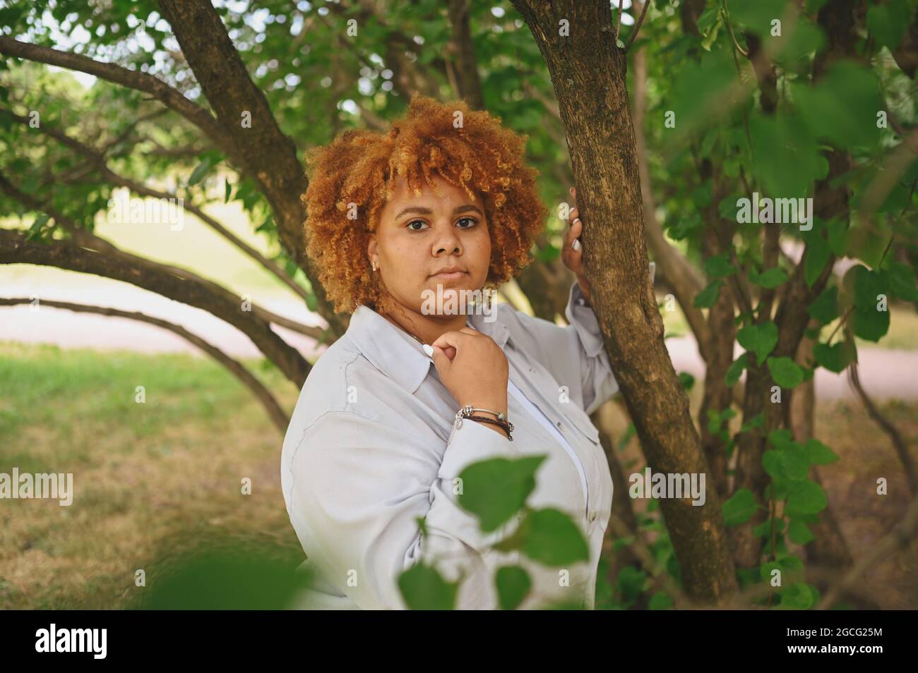 Fashion Portrait von jungen natürlich schönen plus size afroamerikanische Frau in hellen Hosenanzug mit afroroten Haaren posiert in Natur Parkland in grünem Laub. Vielfalt und körperpositives Konzept Stockfoto