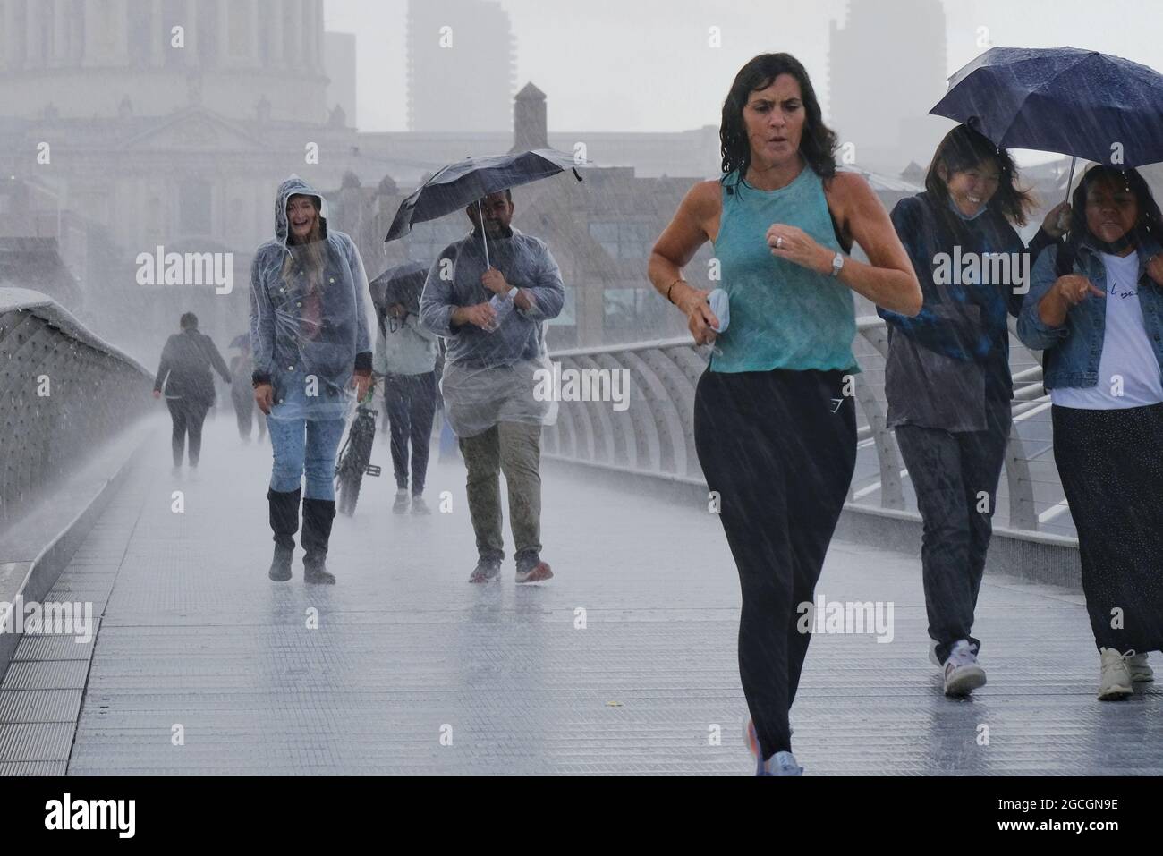 London, Großbritannien. Menschen in feuchter Kleidung, die Regenschirme tragen, und ein Jogger, der vom plötzlichen Regen erwischt wird, eilen entlang der Millennium Bridge. Stockfoto