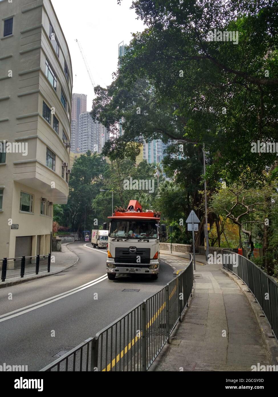 Eine Straße und ein alter Bürgersteig in Hongkong mit einem großen Reinigungs- oder Umzugswagen neben einem glänzenden, modernen Gebäude und üppig grünen Bäumen. Stockfoto