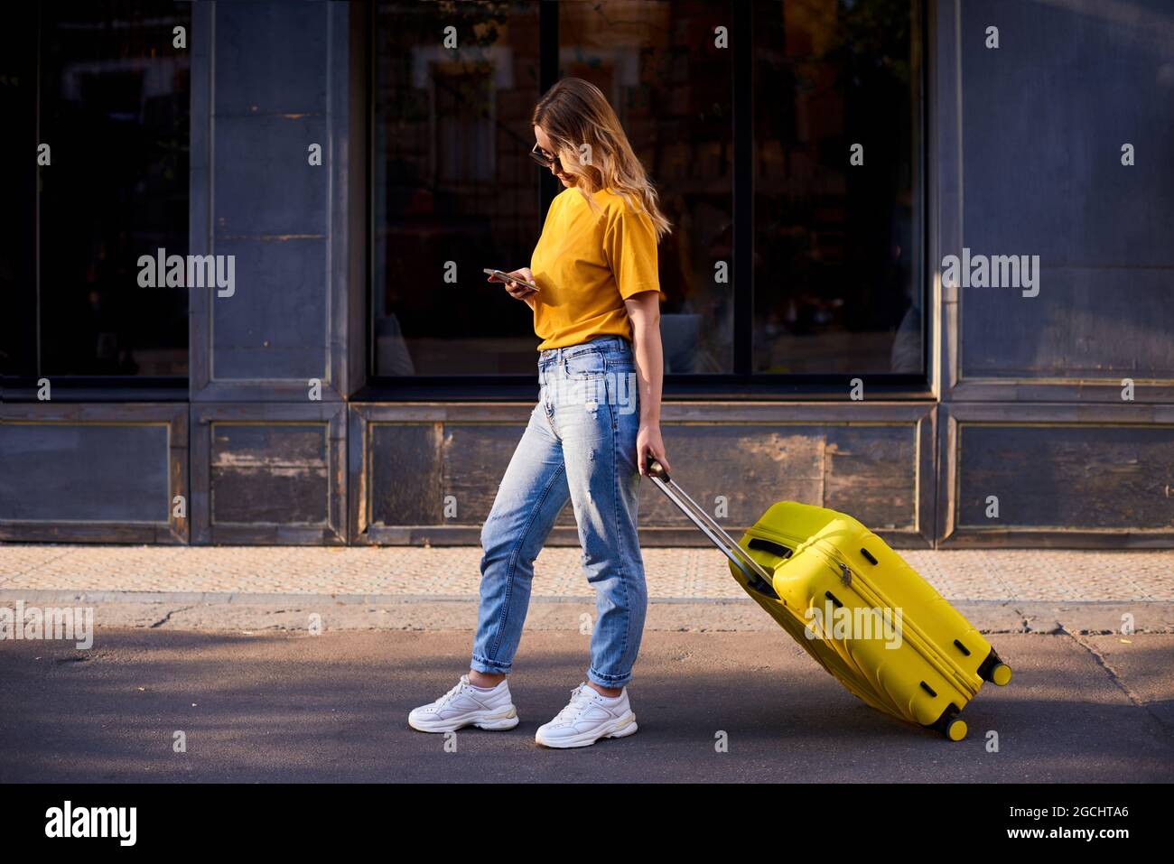 Mädchen Tourist halten Handy in den Händen, gehen durch die Stadt mit gelben Koffer und sehen richtig aus Stockfoto
