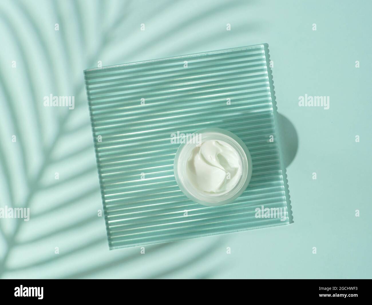 Kosmetische Creme oder Moustirizer auf transparenter gerippter Acrylplatte mit tropischen Palmenblättern Schatten auf blauem Hintergrund. Offenes rundes Glasgefäß mit ästhetischer Wirbelcreme. Flachlagerung von oben. Speicherplatz kopieren Stockfoto