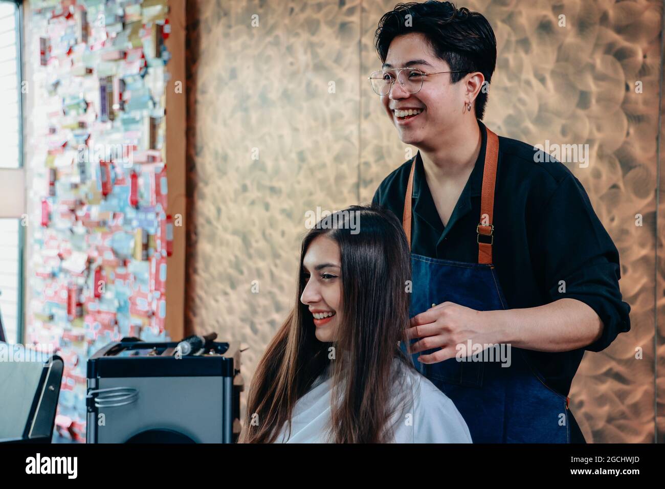Porträt eines männlichen asiatischen Friseurs, der eine Schürze trägt und konzentriert arbeitet, während er in einem modernen Friseurladen hübsche junge Frauenhaare trocknet Stockfoto