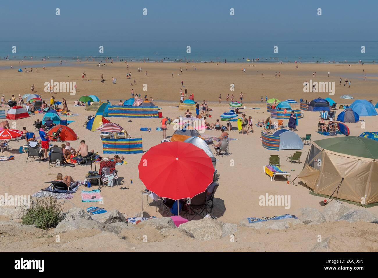 Ein lebhafter roter Sonnenschirm, der von Urlaubern an einem belebten Fistral Beach in den Sommerferien in Newquay in Cornwall verwendet wird. Stockfoto