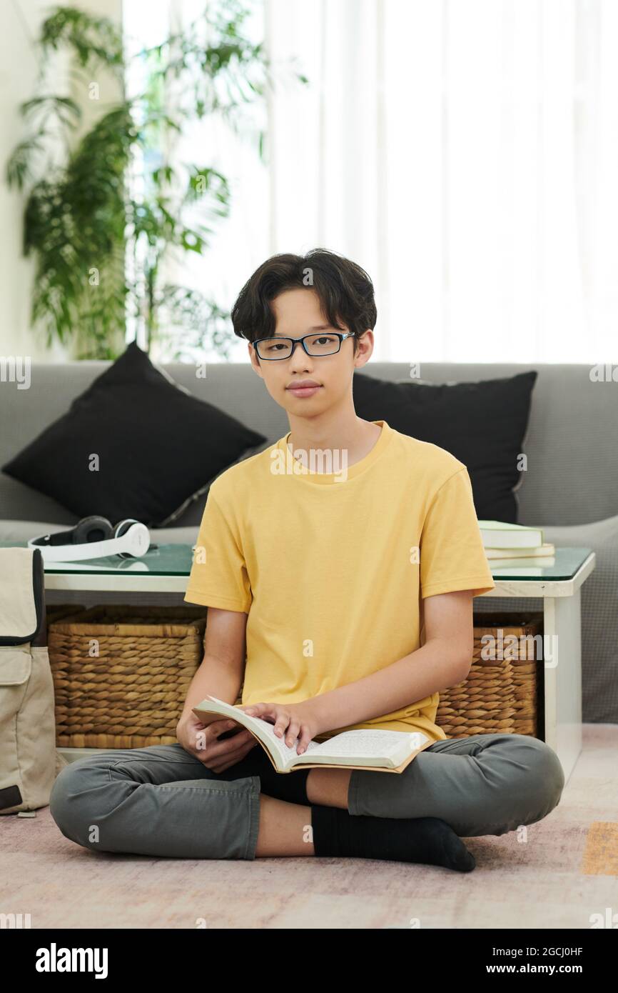 Intelligenter Teenager-Junge mit Brille sitzt zu Hause auf dem Boden und liest ein Buch Stockfoto