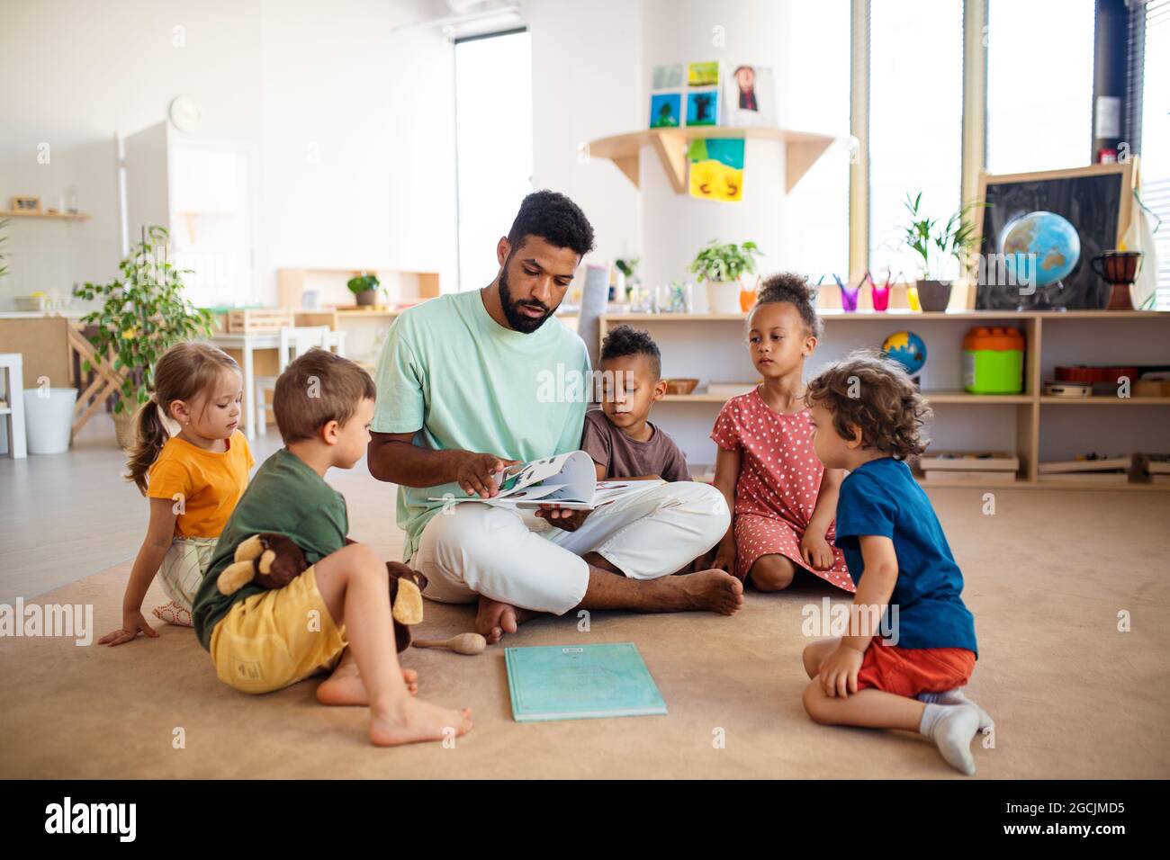 Gruppe von kleinen Kindergarten-Kinder mit Mann Lehrer sitzen auf dem Boden drinnen im Klassenzimmer, mit Unterricht. Stockfoto
